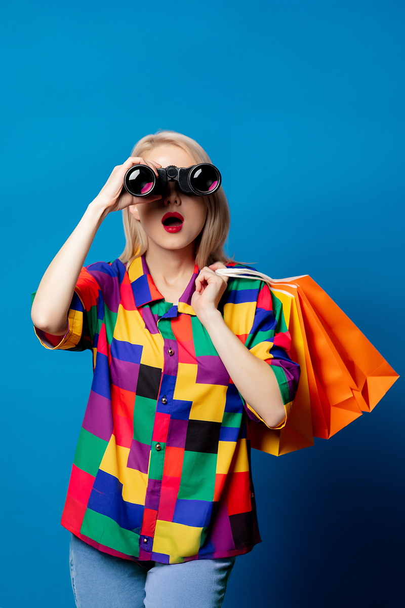 Blonde woman wearing a colorful shirt holding orange shopping bags and looking through binoculars against a blue background, expressing search and discovery.